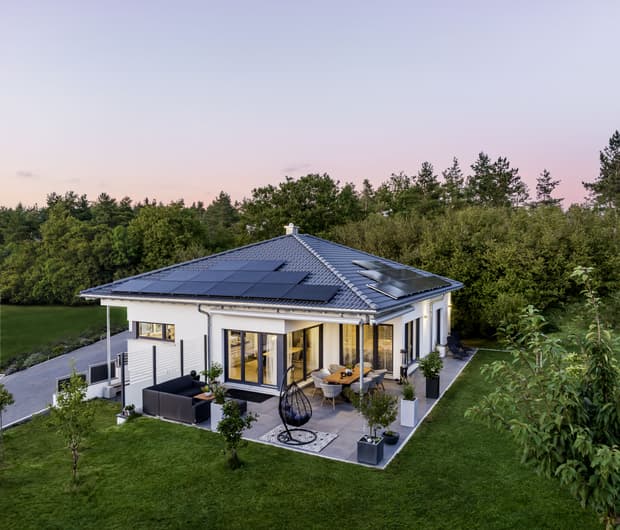 Moderner weißer Bungalow mit grauem Dach und Solarpaneelen umgeben von grünem Garten und Terrasse mit Sitzbereich und Esstisch