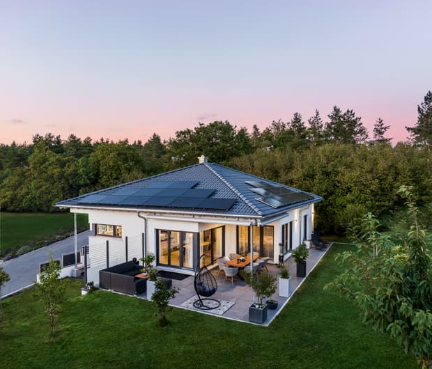 Moderner Bungalow mit Walmdach, beleuchteter Terrasse und Garten in der Abenddämmerung