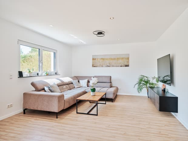 Helles, modern eingerichtetes Wohnzimmer mit großer Eckcouch, Holzoptik-Boden, TV-Wand und großzügigem Fenster