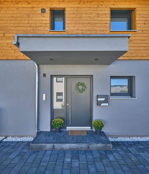 Moderner Hauseingang, graue Haustür mit Seitenteil, Vordach, graue Putzfassade und Holzverkleidung im Obergeschoss