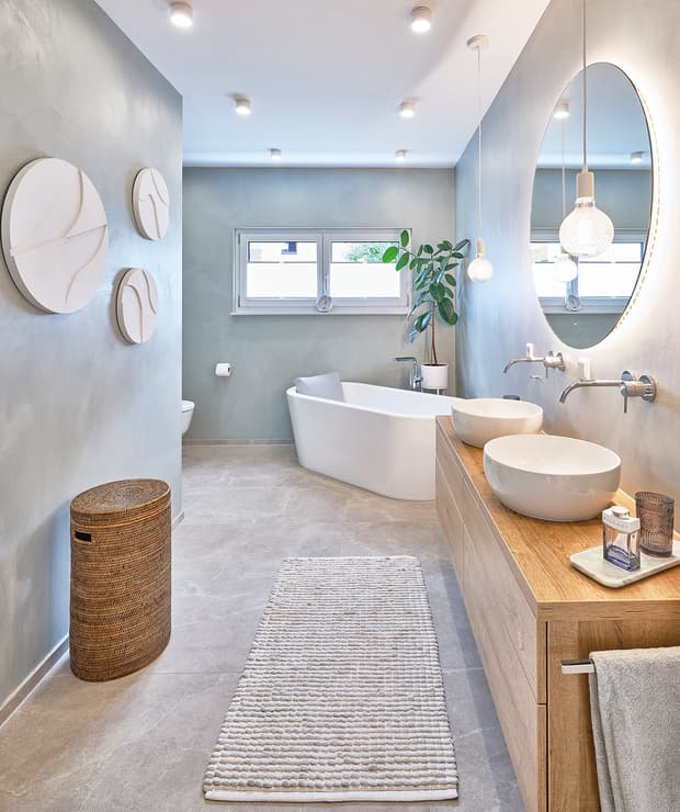 Modernes Badezimmer mit freistehender Badewanne, zwei weißen Aufsatzwaschbecken, Holzmöbeln, runden Wandbildern und Naturkosmetik