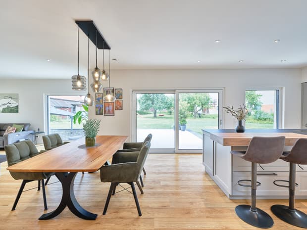 Moderner Essbereich mit Holz­tisch, grünen Polsterstühlen, Kücheninsel und großen Fensterfronten zum Garten