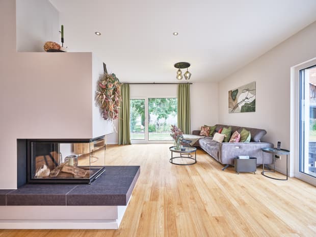 Moderner heller Wohnraum mit Kamin, grauem Sofa, Holzboden und großen Fenstern mit grünem Blick