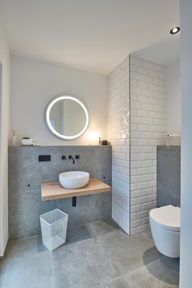 Modernes Badezimmer mit weißem Aufsatzwaschbecken, Holzablage, beleuchtetem Rundspiegel und grauen Fliesen