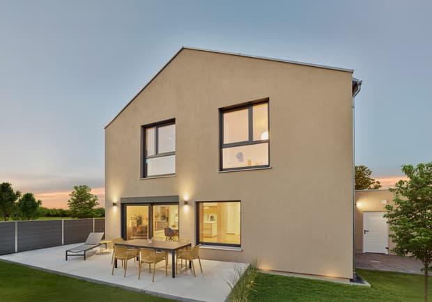 Modernes zweistöckiges Einfamilienhaus mit beigem Putz, großer Terrasse mit Gartenmöbeln und grüner Wiese bei Abendlicht