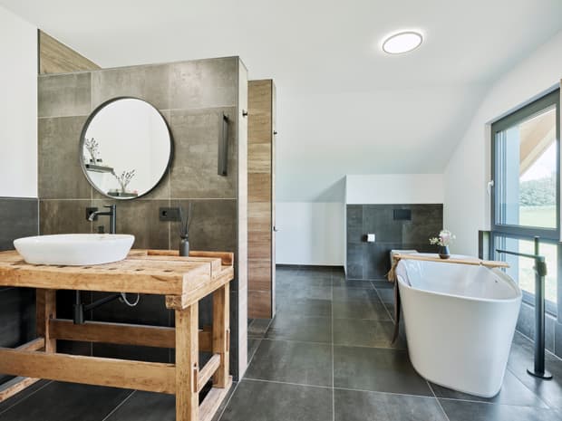 Modernes Badezimmer mit freistehender weißer Badewanne, Holzwaschtisch, rundem Spiegel und großformatigen grauen Fliesen