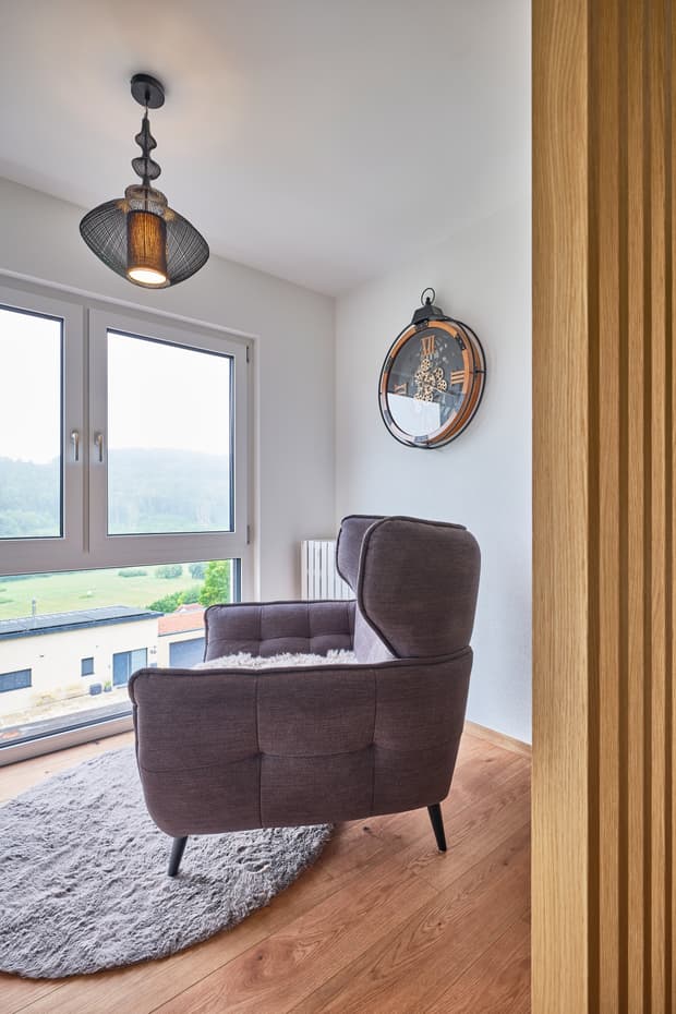Gemütlicher dunkler Stoffsessel auf rundem grauen Teppich vor Fenster mit Ausblick und dekorativer Wanduhr aus Holz und Metall