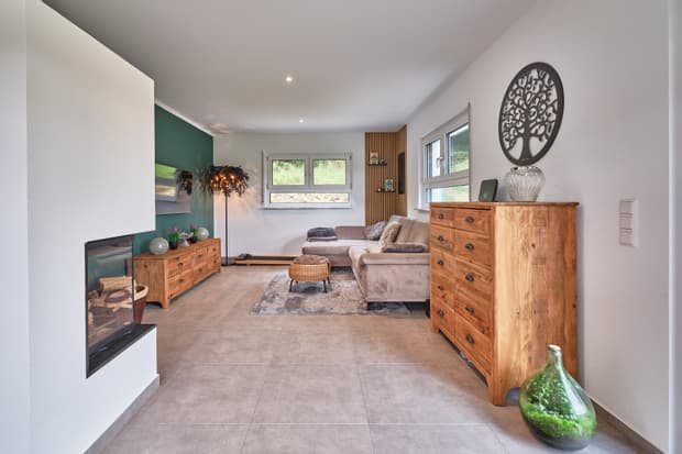 Modern eingerichtetes Wohnzimmer mit grauem Sofa, Holzmöbeln, Kamin und großen Fenstern mit Blick ins Grüne