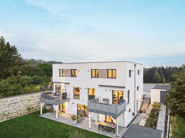 Modernes weißes Doppelhaus mit großen Fenstern und Balkonen umgeben von grünem Garten und Bäumen bei Abendlicht