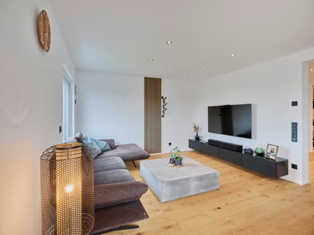 Moderner Wohnraum mit braunem Sofa, großem quadratischem Betontisch, schwarzer Wandhalterung und Holzparkettboden