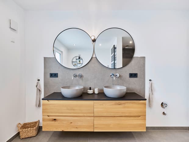 Modernes Badezimmer mit Doppelwaschbecken aus grauem Stein und Holzunterschrank vor grauer Wand mit zwei runden Spiegeln