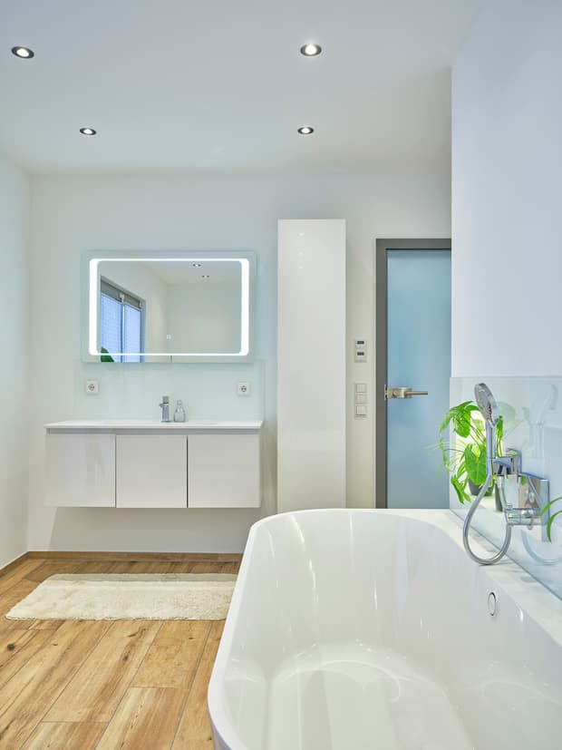 Modernes Badezimmer mit frei stehender Badewanne, Holzoptik-Fliesen, beleuchtetem Spiegel und grünem Pflanzenakzent