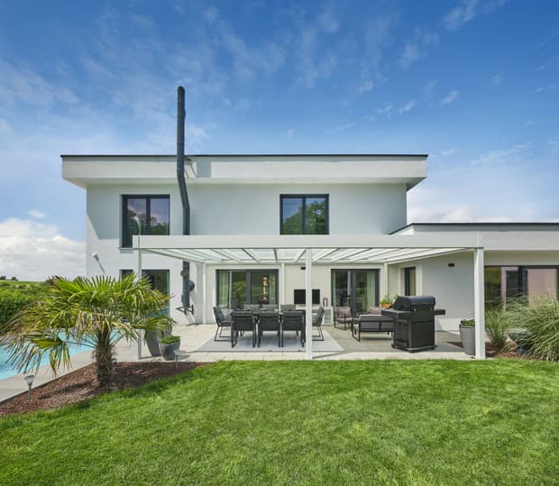 Moderner weißer zweistöckiger Bungalow mit überdachter Terrasse, Gartenmöbeln, Grill und grünem Rasen bei blauem Himmel