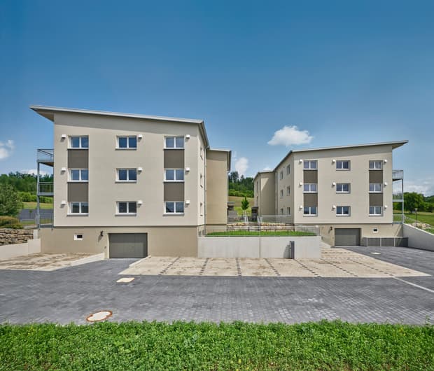 Zwei moderne Wohngebäude mit hellem Putz, grauen Akzenten und Garagen in ländlicher Umgebung bei klarem Himmel