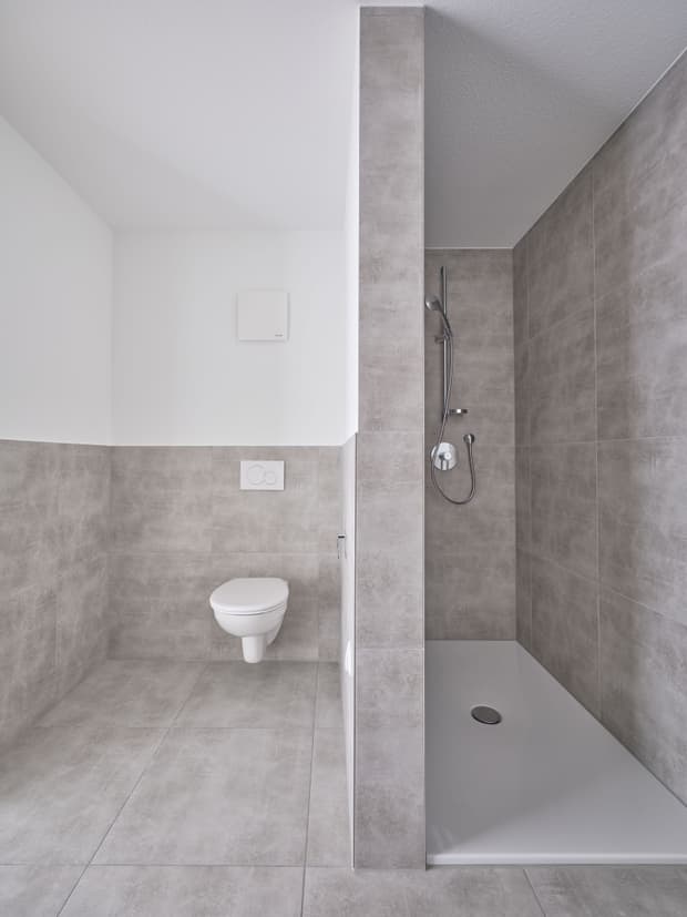 Modernes Badezimmer mit grauen Fliesen, weißer wandhängender Toilette und offener Dusche mit Duschkopf und Duscharmatur