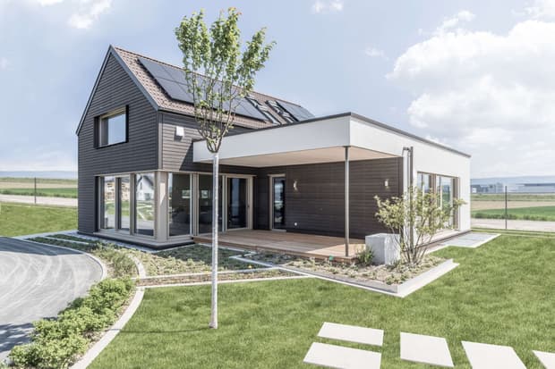 Modernes Musterhaus mit Holzfassade, Satteldach, Solarpanelen, überdachter Terrasse und gepflegtem Garten.