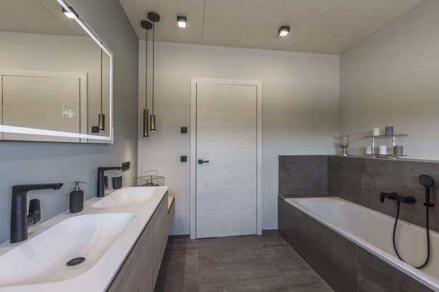 Modernes Badezimmer im Musterhaus mit Doppelwaschbecken, Badewanne, grauen Fliesen und stilvoller Beleuchtung.