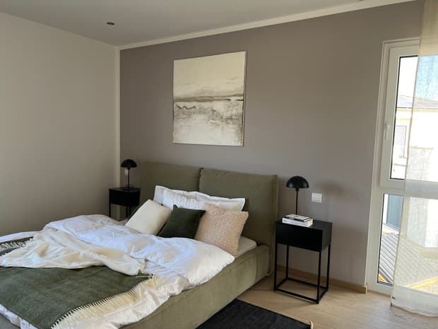Modernes Schlafzimmer im Musterhaus mit grauer Akzentwand, Doppelbett, Kunstbild, Nachttischen und Tageslicht.
