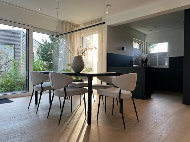 Helles Esszimmer im modernen Musterhaus mit rundem Tisch, Designerstühlen, großer Fensterfront und offener Küche.