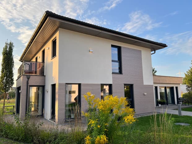 Modernes zweistöckiges Musterhaus mit Flachdach, heller Fassade, großen Fenstern, Balkon und Garten im Abendlicht.