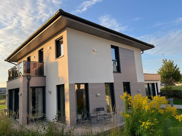 Modernes zweistöckiges Musterhaus mit Flachdach, heller Fassade, Balkon, großen Fenstern und Garten im Abendlicht.