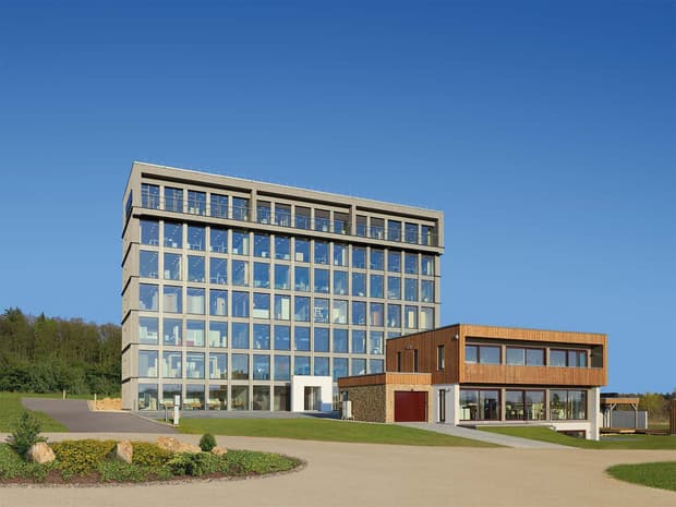 Modernes mehrstöckiges Bürogebäude mit großer Glasfassade und rechteckigem Rasterdesign neben einem niedrigeren Gebäude mit Holzverkleidung. Die Gebäude stehen in einer offenen Landschaft unter wolkenlosem blauem Himmel.