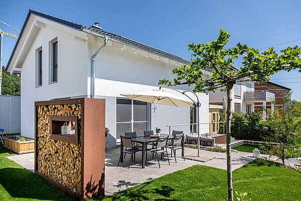 Moderner weißer Bungalow mit Terrasse, Esstisch, Sonnenschirm und dekorativem Holzlager im Garten