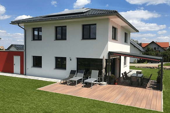 Modernes weißes Einfamilienhaus mit Sonnenterrasse, Gartenmöbeln und großer Rasenfläche unter blauem Himmel