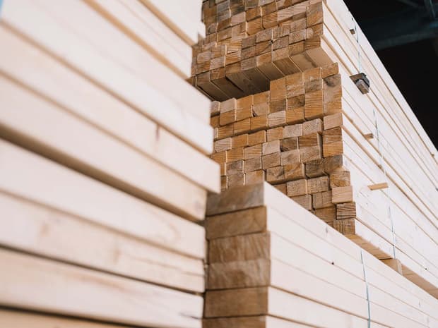 Mehrere Stapel von Holzlatten und -balken liegen dicht nebeneinander, teils ordentlich, teils ungleichmäßig gestapelt. Die Perspektive zeigt die Schnittflächen und Maserung des Holzes.