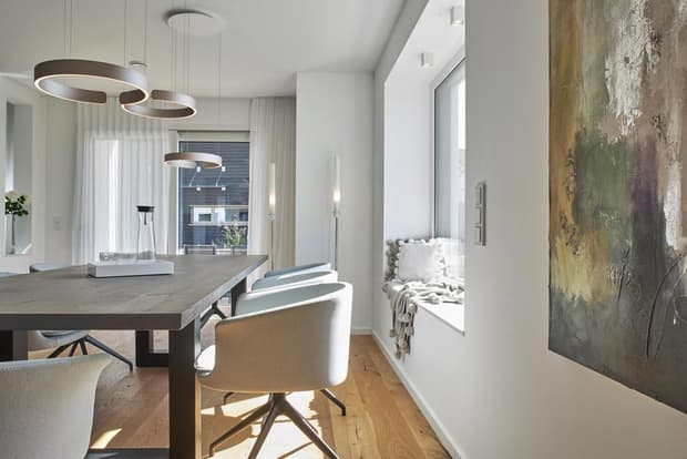 Helles Esszimmer im modernen Musterhaus mit Holzboden, großem Esstisch, Designerlampen und gemütlicher Fensterbank.