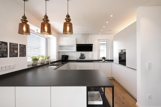 Moderne Küche im Musterhaus mit schwarzer Arbeitsplatte, weißen Schränken, Kupferlampen und hellem Holzboden.