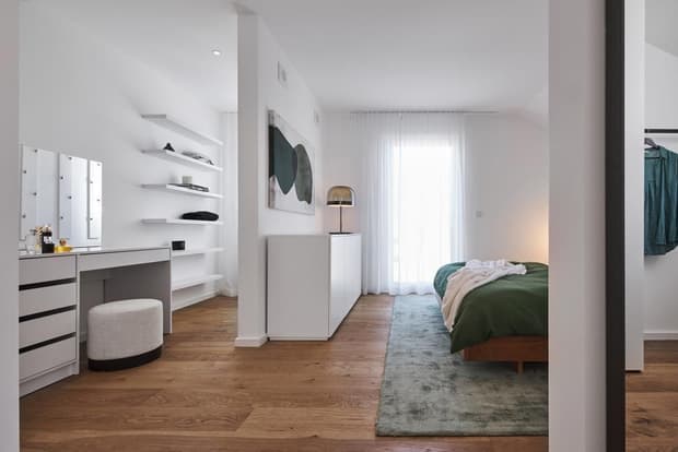 Helles Schlafzimmer im Musterhaus mit Holzboden, grünem Bett, Schminktisch, Regalwand und bodentiefem Fenster.