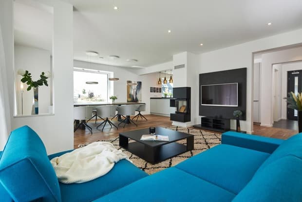 Modernes Musterhaus-Wohnzimmer mit blauer Couch, Kamin, großem Fernseher, Esstisch und heller, offener Raumgestaltung.