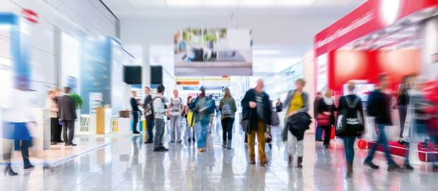 Verschwommene Menschenmenge in heller Messehalle mit Ausstellungsständen und glänzendem Boden