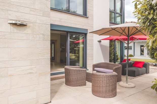 Terrassenmöbel aus Geflecht mit grauen Polstern unter hellem Sonnenschirm an modernem Haus mit Sandsteinfassade