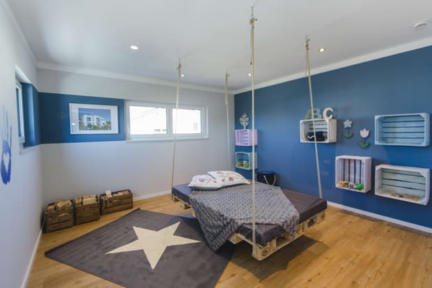 Modernes Kinderzimmer im Musterhaus mit hängendem Bett, blauer Wand, Holzboden und dekorativen Wandregalen.