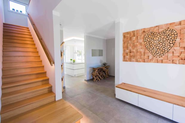 Heller Eingangsbereich im modernen Musterhaus mit Holztreppe, Wandkunst in Herzform und offener Küche.