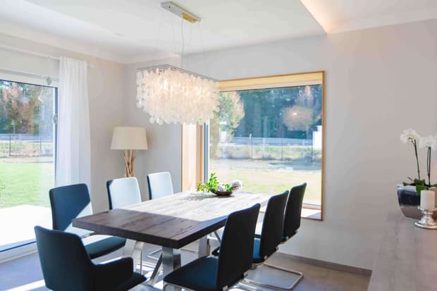 Helles Esszimmer im modernen Musterhaus mit Holztisch, blauen Stühlen, großer Fensterfront und Designlampe.
