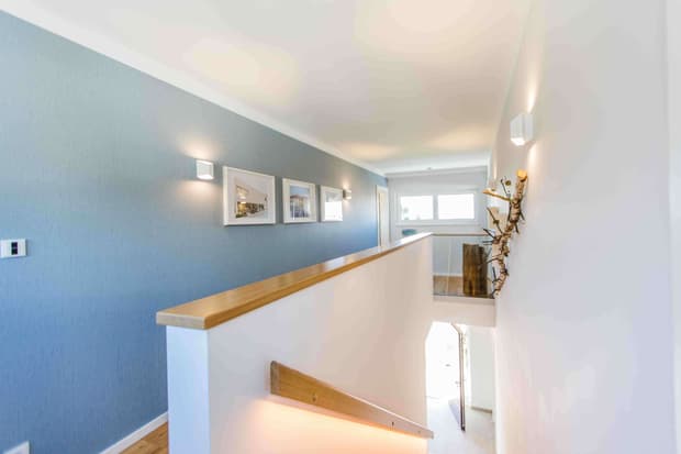 Heller Flur im modernen Musterhaus mit blauer Akzentwand, Holzgeländer, Wandbeleuchtung und stilvollen Bildern.