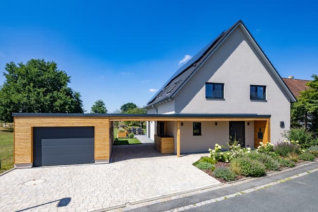 Modernes Einfamilienhaus mit weißer Fassade, Holz-Carport und Garage, Solarzellen auf dem Dach, Garten mit Blumen und Sträuchern