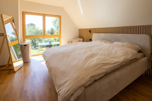 Schlafzimmer mit großem Bett in hellem Stoff, Holzfußboden, Fenster mit Naturblick, Stehspiegel und Wäschekorb aus Stoff