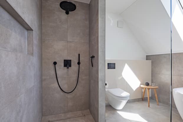 Moderne Badezimmerdusche mit schwarzen Armaturen und matte graue Fliesen neben weißer Toilette und Holzhocker unter Dachschräge