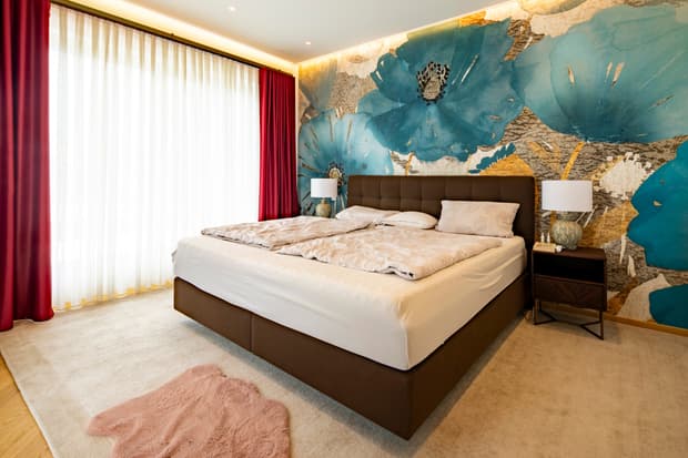 Modernes Schlafzimmer mit braunem Polsterbett, blauer Blumen-Wandtapete, roten Vorhängen und hellbeigem Teppich