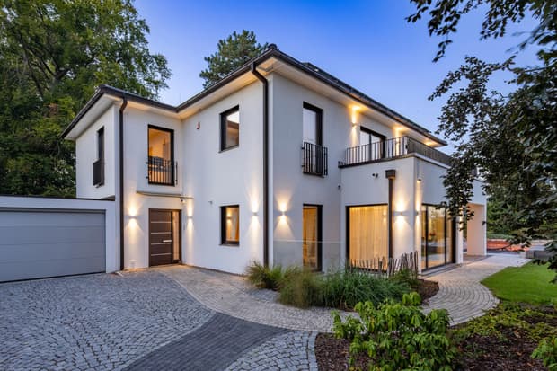 Modernes zweistöckiges Einfamilienhaus mit weißer Fassade, großen Fenstern, beleuchtet bei Dämmerung, umgeben von Garten und Bäumen
