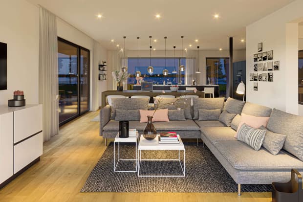 Offenes Wohnzimmer im modernen Musterhaus mit grauer Eckcouch, Holzboden, Esstisch, Küche und stimmungsvoller Beleuchtung.