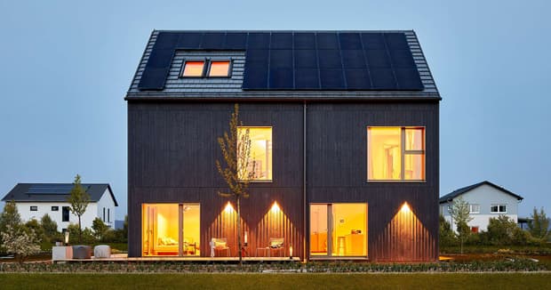 Modernes schwarzes Musterhaus mit Satteldach, großen Fenstern, Solarpanelen und warm beleuchtetem Innenraum bei Abendlicht.