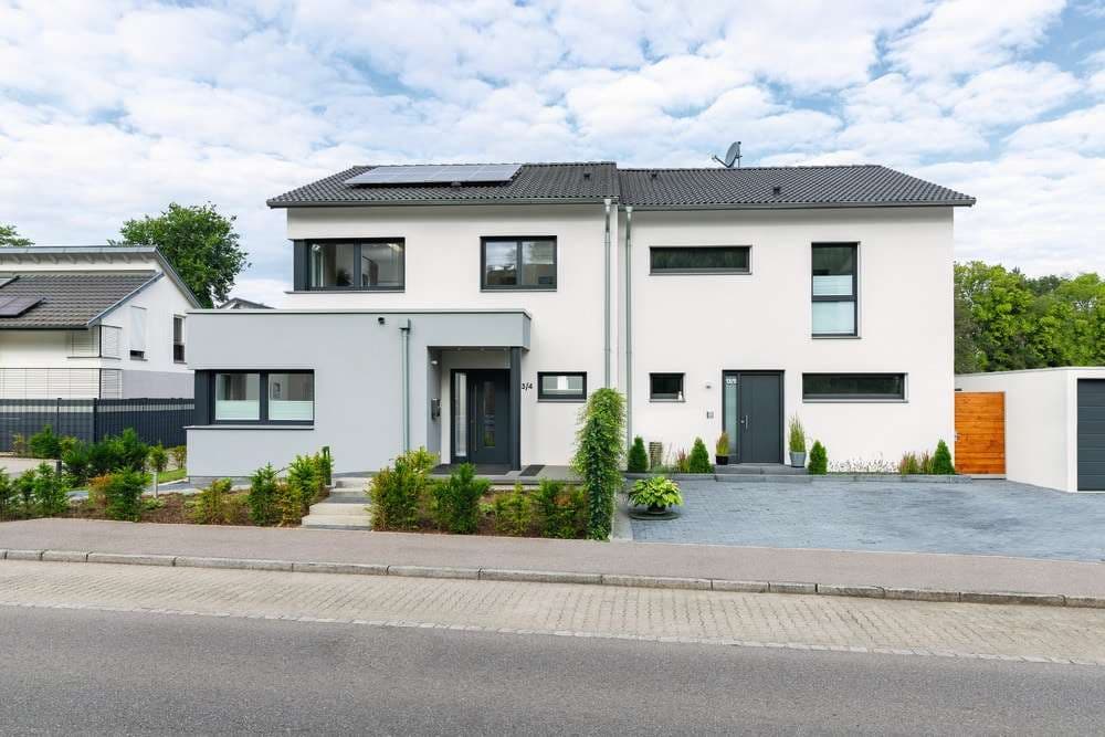 Moderne Doppelhaushälfte mit Flachdach- und Satteldachkombination, weißer Putzfassade, grauen Fensterrahmen und gepflasterter Einfahrt.