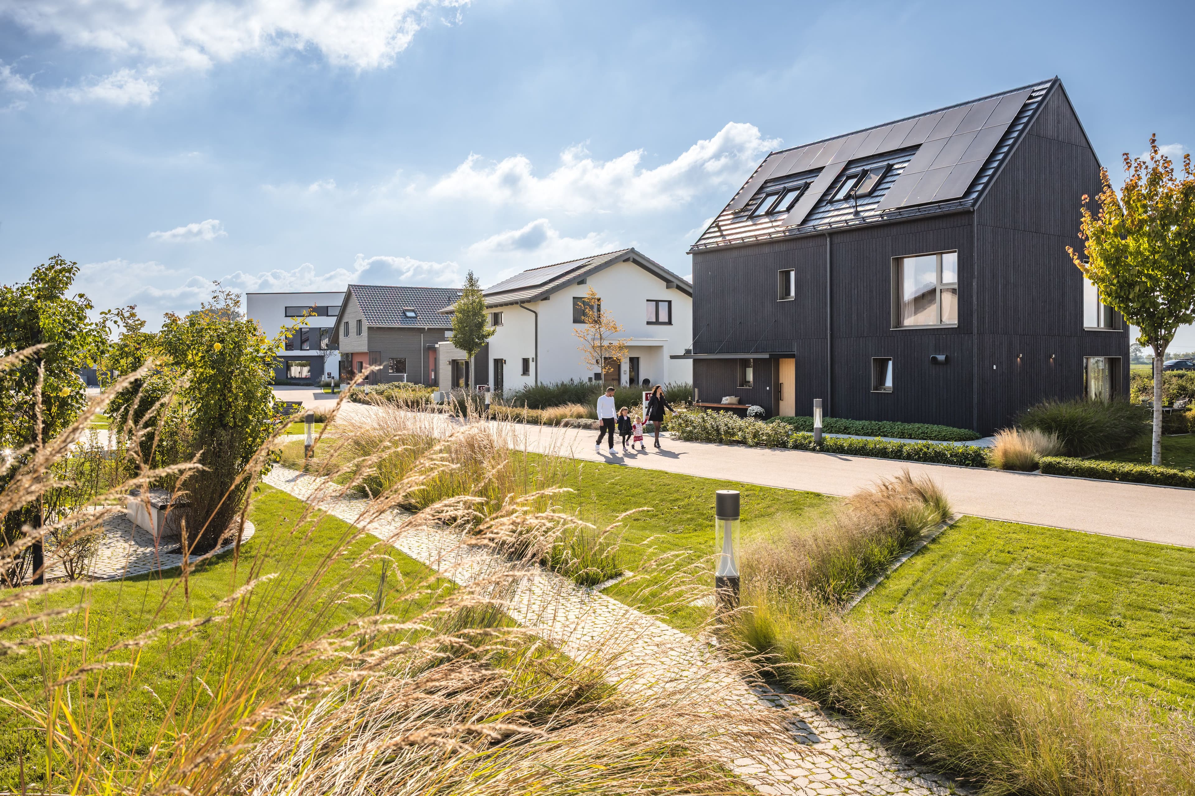 Moderne Fertighaussiedlung mit mehreren Einfamilienhäusern, grünen Vorgärten und Spaziergängern bei sonnigem Wetter.