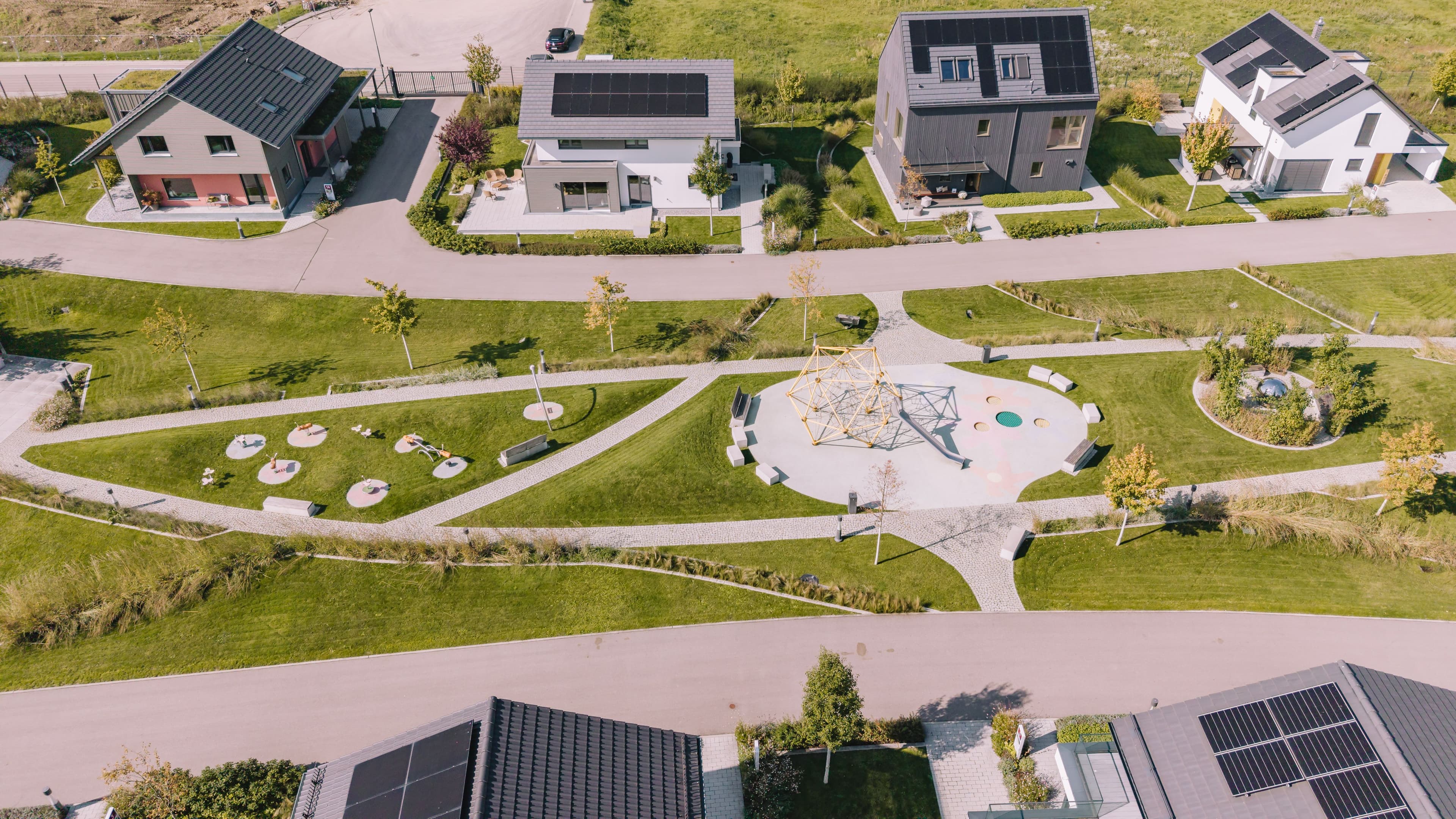 Luftaufnahme einer modernen Wohnsiedlung mit energieeffizienten Fertighäusern, Solardächern, gepflegten Grünflächen und zentralem Spielplatz mit Klettergerüst.