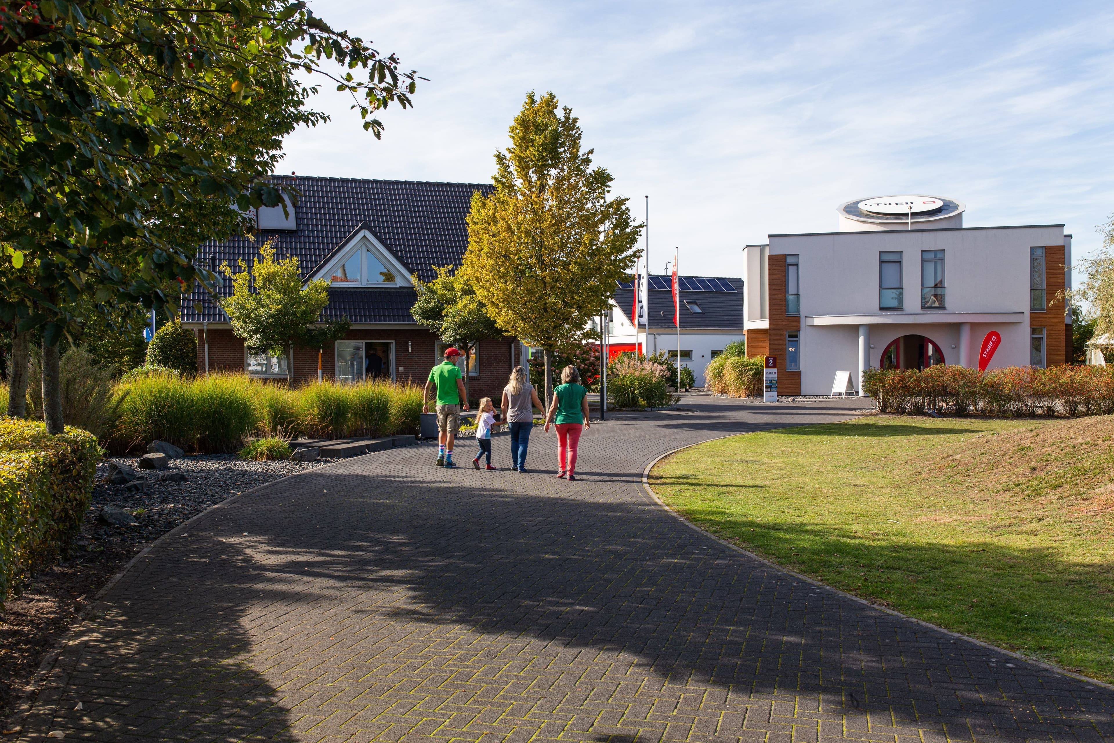Luftaufnahme einer Hausausstellung mit modernen Musterhäusern, großem Parkplatz voller Autos, grüner Umgebung und sonnigem Himmel über der Stadt.